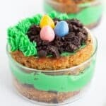 A small glass dish contains a layered dessert with green frosting, crumbled cookies, piped green icing, and three colorful candy eggs on top—one of the cutest Mini Easter Trifles perfect for your collection of Easter desserts.