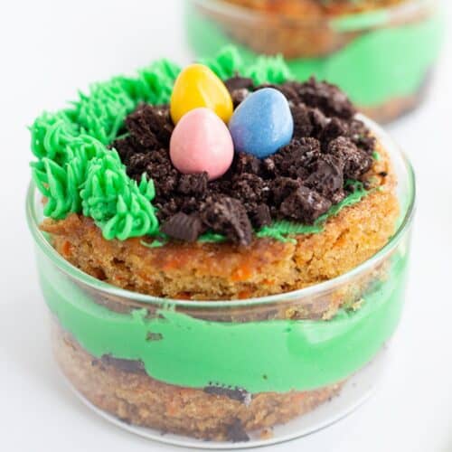 A small glass dish contains a layered dessert with green frosting, crumbled cookies, piped green icing, and three colorful candy eggs on top—one of the cutest Mini Easter Trifles perfect for your collection of Easter desserts.