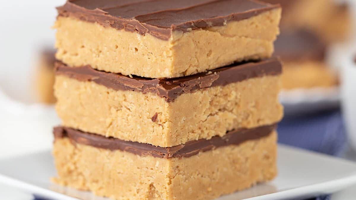 Three thick squares of no bake peanut butter bars topped with chocolate are stacked on a white plate&mdash;perfect for easy desserts or winter desserts.