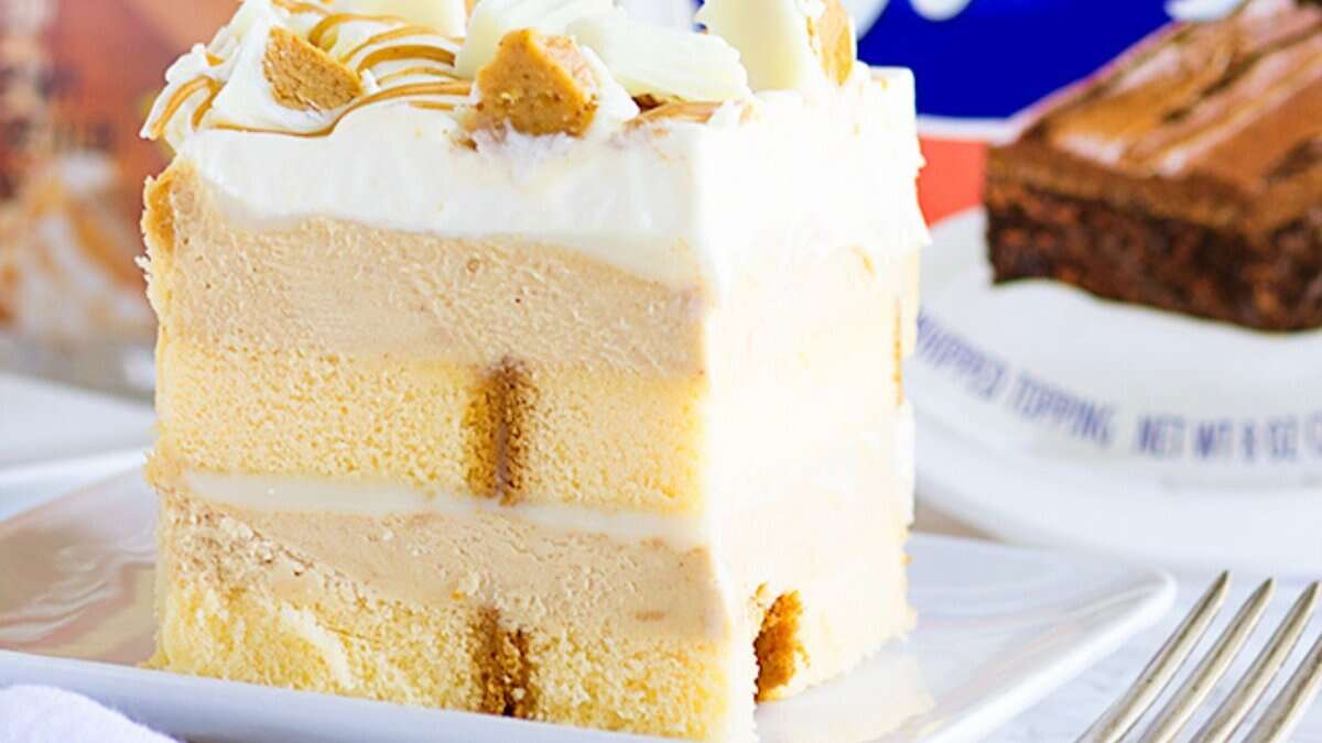 Slice of layered cake with vanilla sponge, creamy filling, whipped topping, and white chocolate pieces on a white plate&mdash;an easy dessert perfect for fans of no bake desserts. Another tempting treat appears in the background.