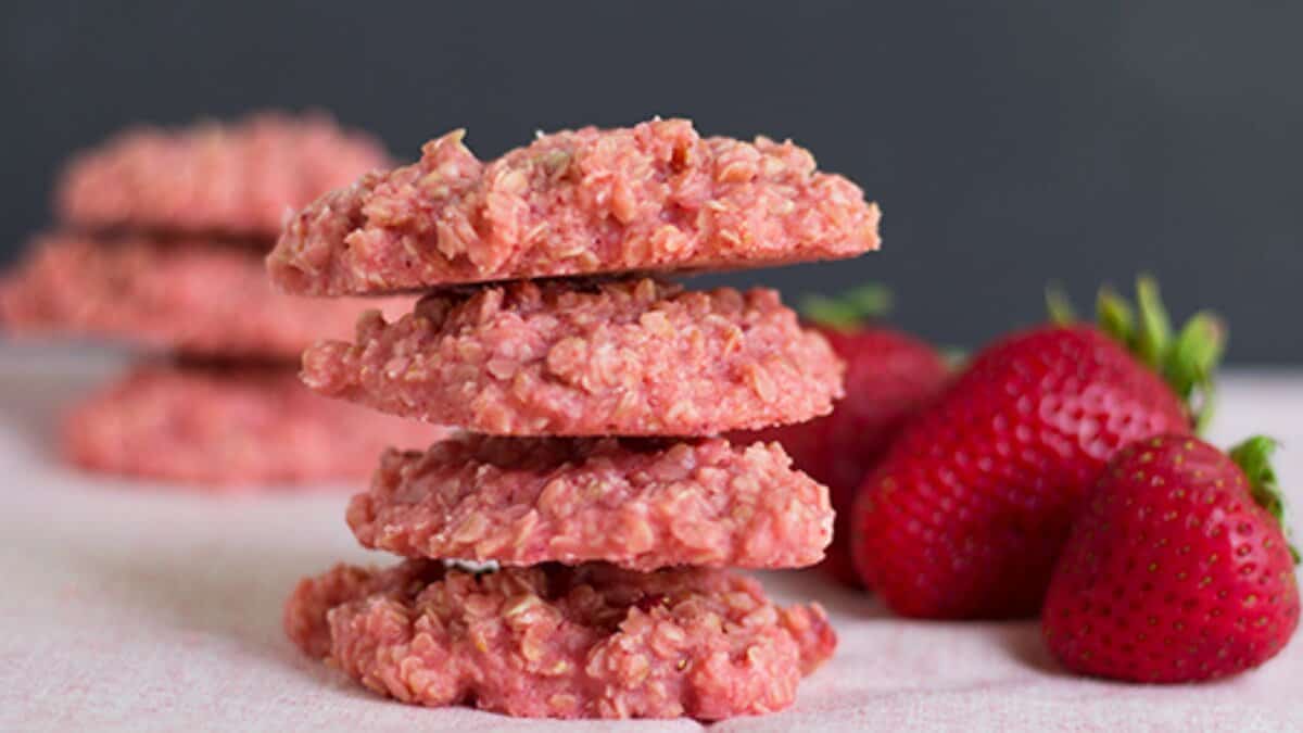 A stack of pink strawberry oatmeal cookies sits next to fresh strawberries, offering a delightful twist on easy desserts, with more cookies blurred in the background.