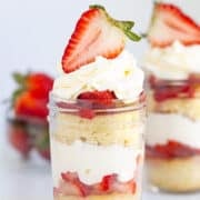 A Strawberry Shortcake In A Jar, featuring layers of sponge cake, whipped cream, and sliced strawberries, topped with a halved strawberry. Another dessert jar and fresh strawberries sit in the background.