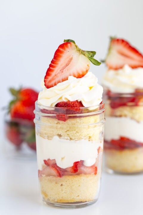 A Strawberry Shortcake In A Jar, featuring layers of sponge cake, whipped cream, and sliced strawberries, topped with a halved strawberry. Another dessert jar and fresh strawberries sit in the background.