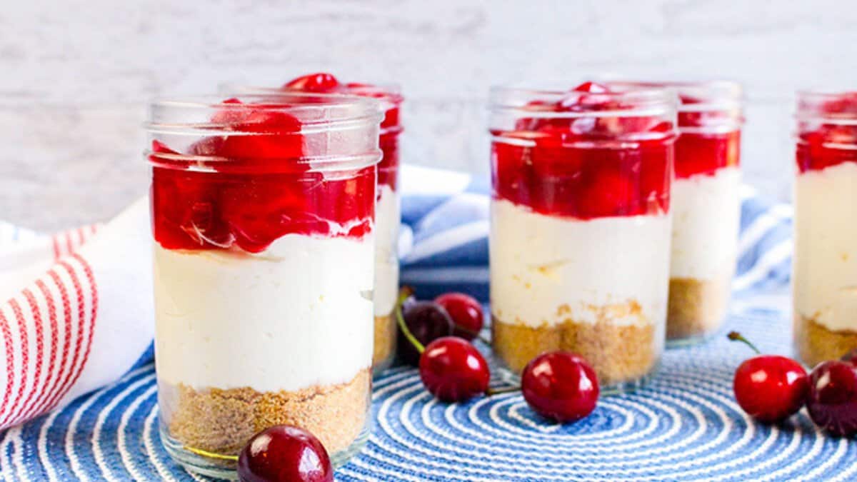 Five glass jars filled with layered cherry cheesecake parfaits on a blue and white placemat, garnished with cherries and surrounded by fresh cherries&mdash;perfect no bake desserts for any occasion.