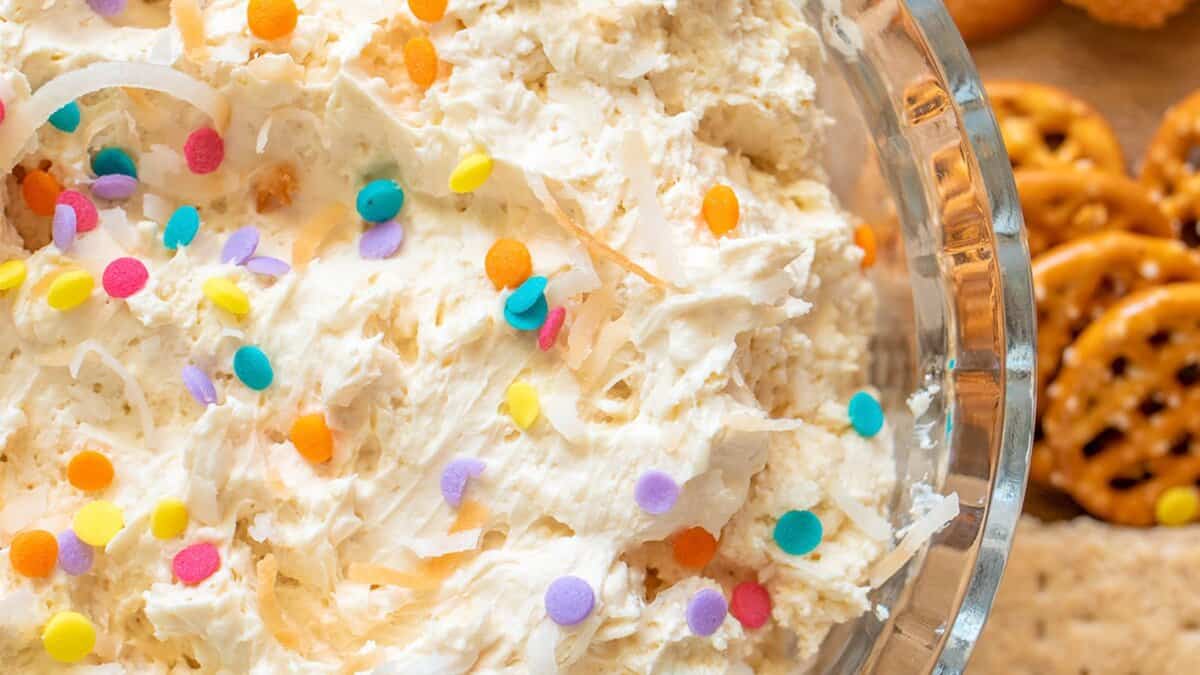 A close-up of a glass bowl filled with a creamy no bake dessert dip topped with colorful round sprinkles, next to pretzels and crackers.