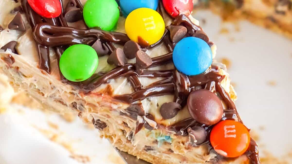A slice of creamy, no bake dessert topped with colorful candy-coated chocolates, mini chocolate chips, and a drizzle of chocolate syrup&mdash;perfect for easy desserts or festive winter desserts.