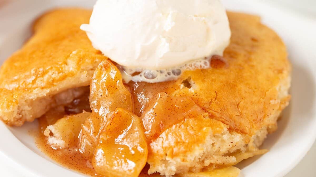A serving of apple cobbler, a classic dessert, topped with a scoop of vanilla ice cream on a white plate.