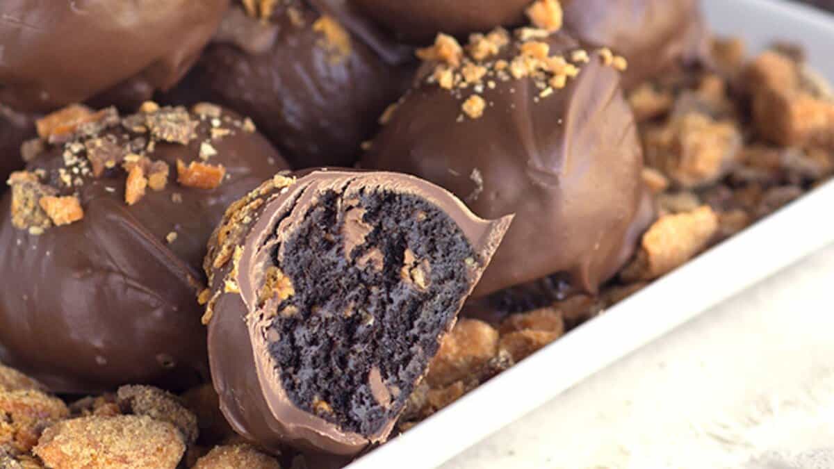 Close-up of chocolate-coated cake balls, one cut open to reveal a dark, moist interior, topped and surrounded by cookie crumbs on a white tray&mdash;perfect for those seeking simple dessert recipes or easy desserts.