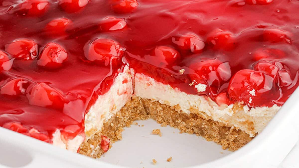 A close-up of a traditional dessert with a graham cracker crust, creamy filling, and cherry topping in a white dish with a portion removed.