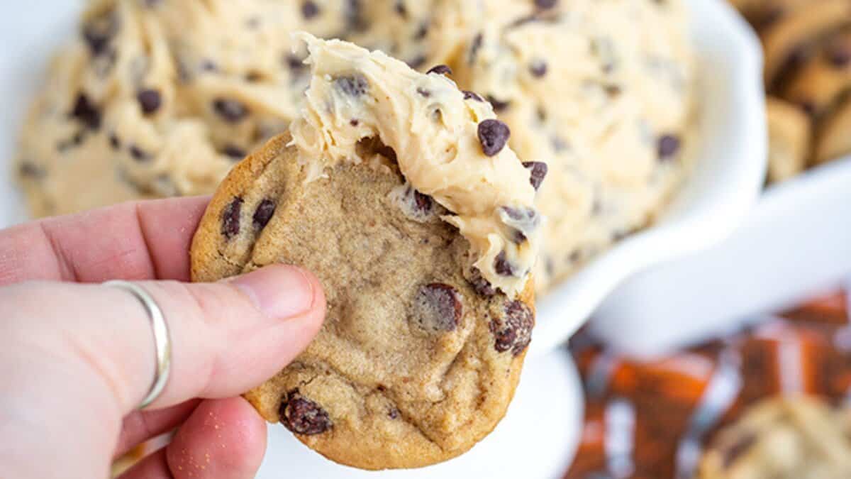 A hand holding a chocolate chip cookie topped with a scoop of cookie dough, with a bowl of dough in the background&mdash;perfect for those seeking stress-free desserts.