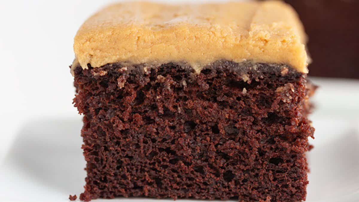 A close-up of a slice of chocolate cake with a thick layer of light brown frosting on top, shown on a white surface&mdash;an irresistible treat among classic desserts.