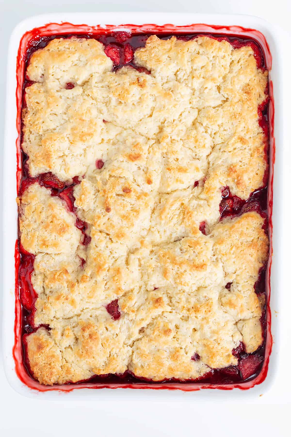 A baked strawberry cobbler in a white rectangular dish, featuring a golden-brown biscuit topping with visible red fruit filling along the edges&mdash;an irresistible strawberry dessert perfect for any cobbler recipe collection.