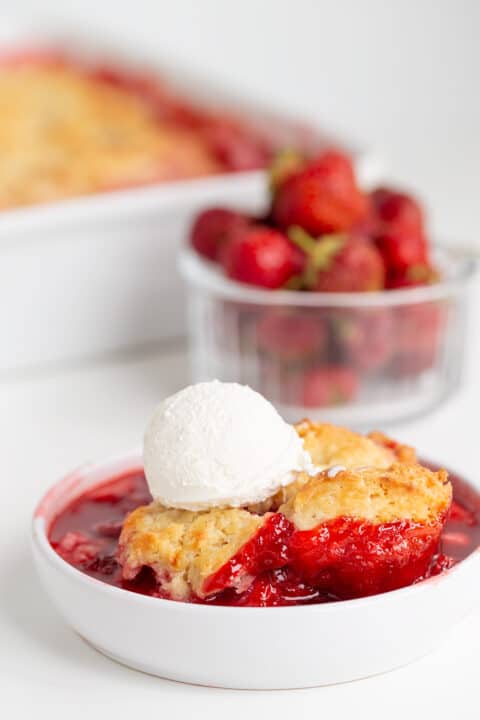 A bowl of Strawberry Cobbler topped with a scoop of vanilla ice cream, surrounded by fresh strawberries and a baking dish, makes for the perfect strawberry dessert.