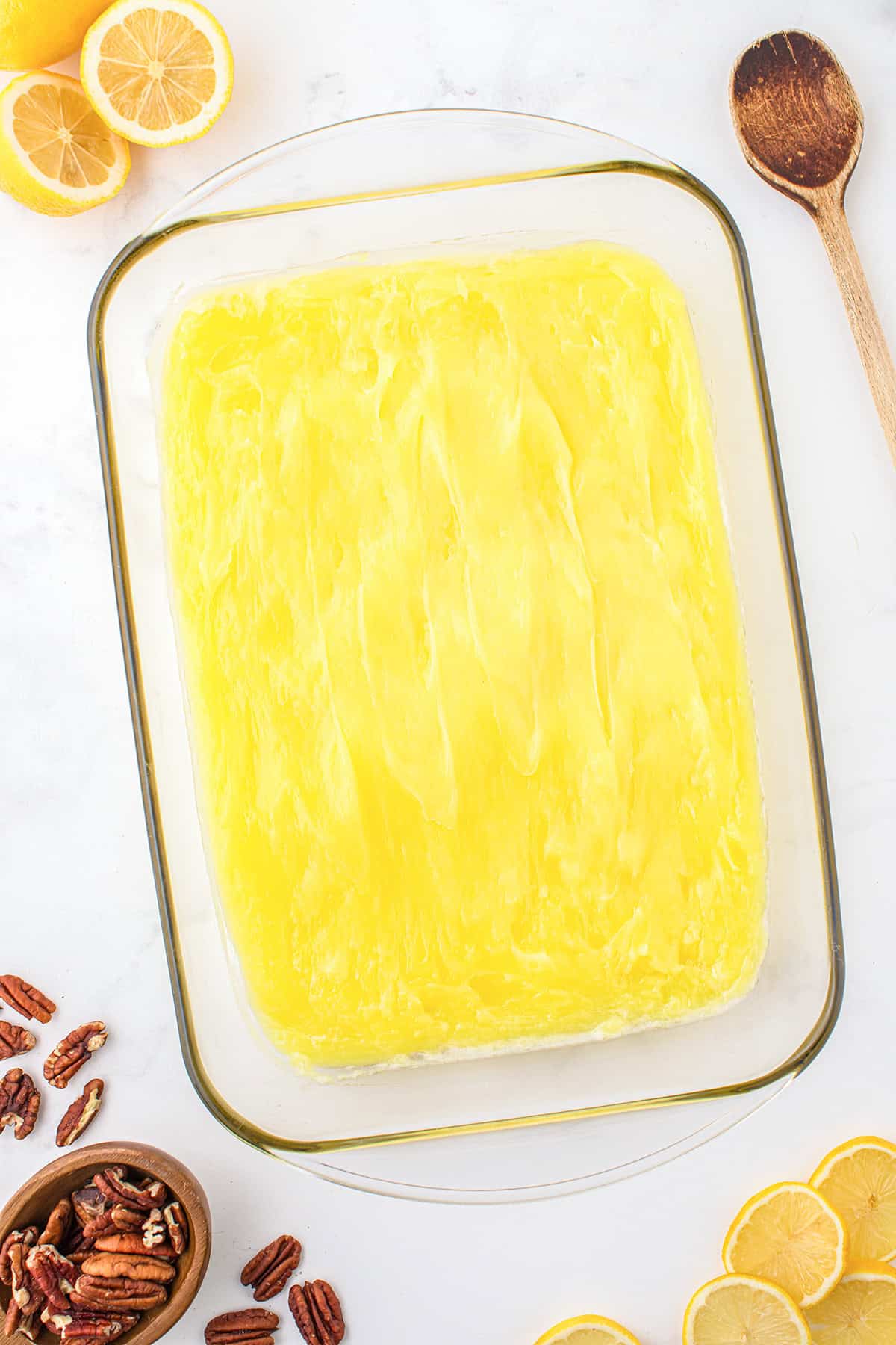 A glass baking dish filled with a smooth layer of yellow lemon gelatin dessert, surrounded by lemon slices, pecans, and a wooden spoon on a white surface—perfect alongside your favorite lemon dump cake.