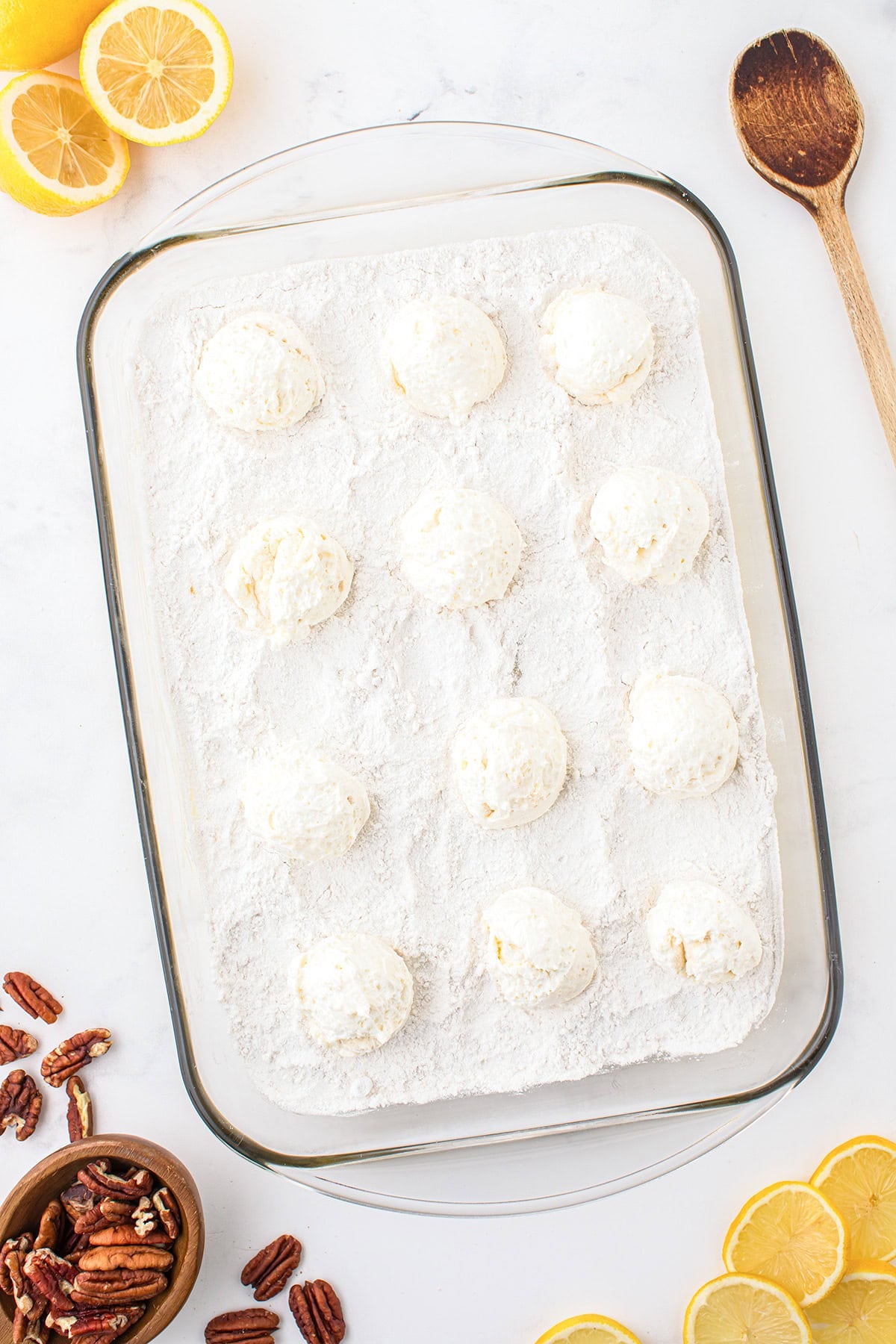 Glass baking dish with cookie dough balls coated in flour, surrounded by lemon slices, pecans, a wooden spoon, and a small bowl of pecans—perfect for creating a zesty Lemon Cake or an easy Dump Cake on a bright white surface.