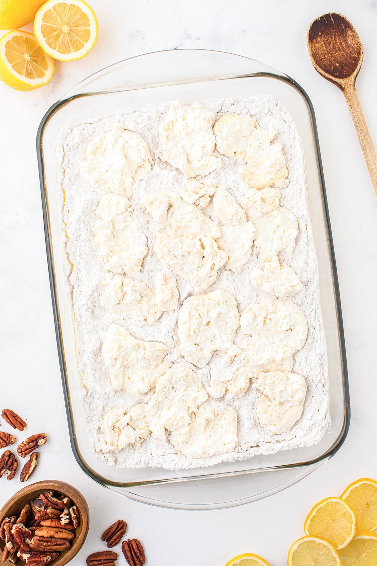 A glass baking dish filled with a flour mixture and dollops of dough, surrounded by lemon halves, pecans, a wooden spoon, and a sliced lemon on a white surface—perfect for making a tangy lemon dump cake or your favorite lemon dessert.