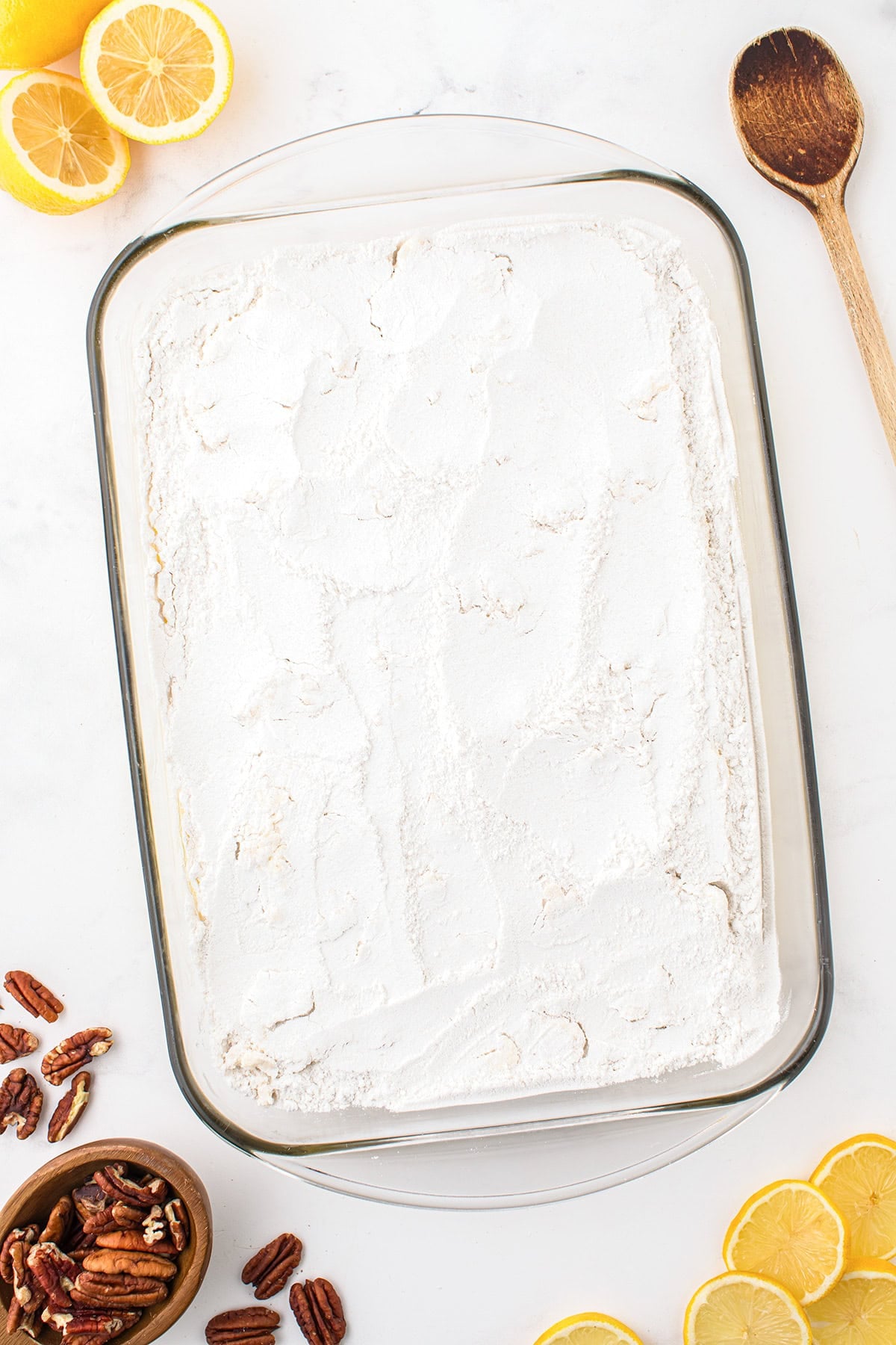 A glass baking dish filled with a white powdered topping, surrounded by lemon halves, pecans, a bowl of pecans, and a wooden spoon on a white surface—perfect for serving as a Lemon Dump Cake.