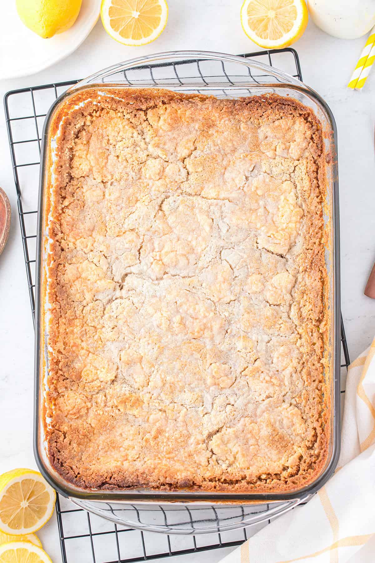 A rectangular glass baking dish filled with a golden-brown, cracked lemon dump cake sits on a cooling rack, surrounded by sliced lemons.