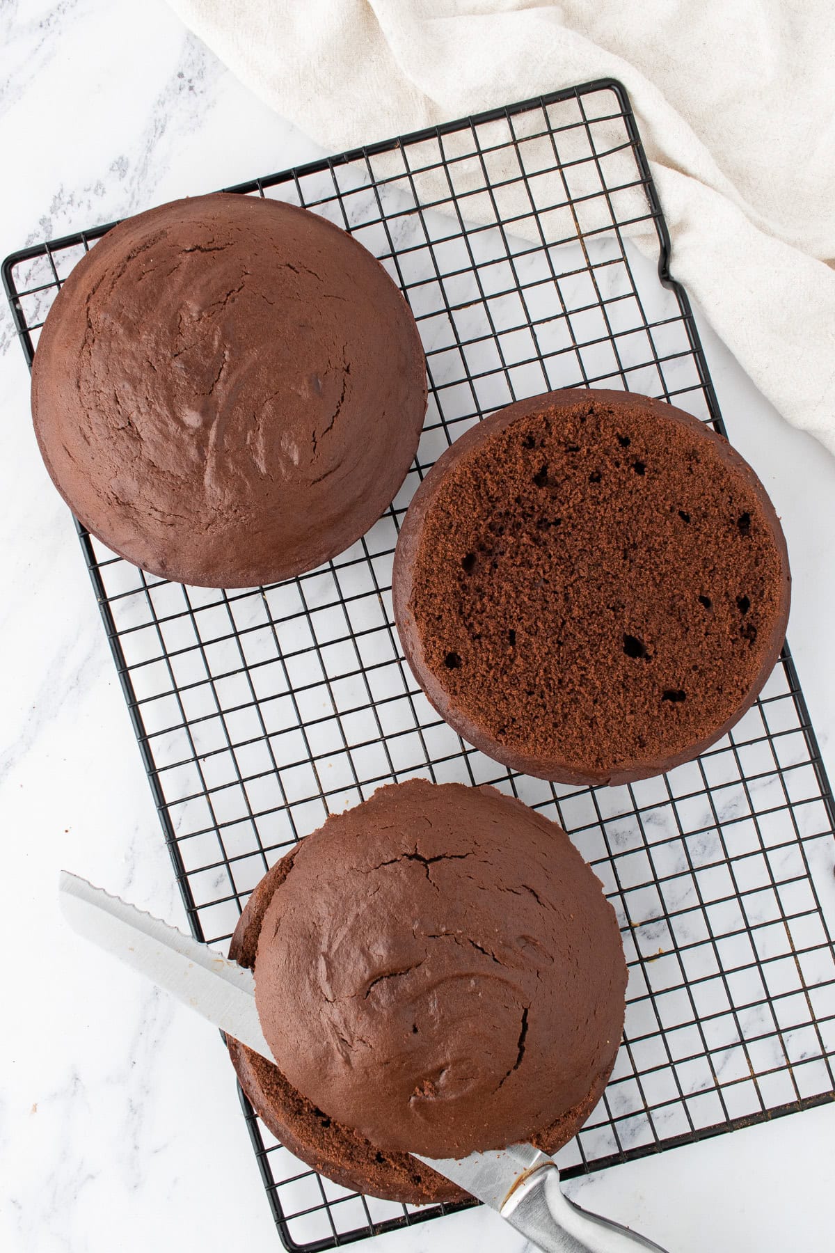 Three round Peanut Butter Chocolate Cake layers cooling on a black wire rack, with a bread knife slicing one layer; white marble surface underneath.