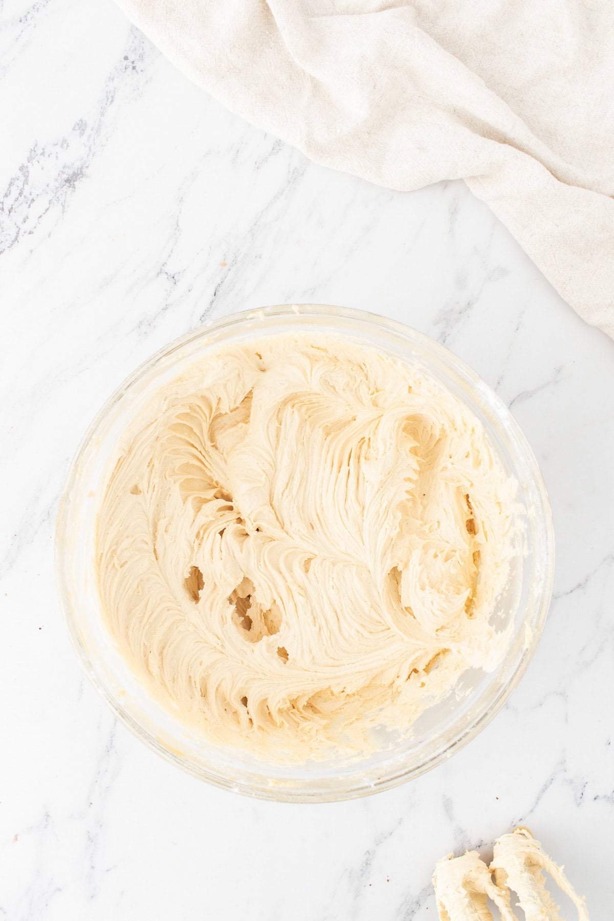 A glass bowl filled with creamy, peanut butter batter sits on a white marble countertop next to a beige cloth.