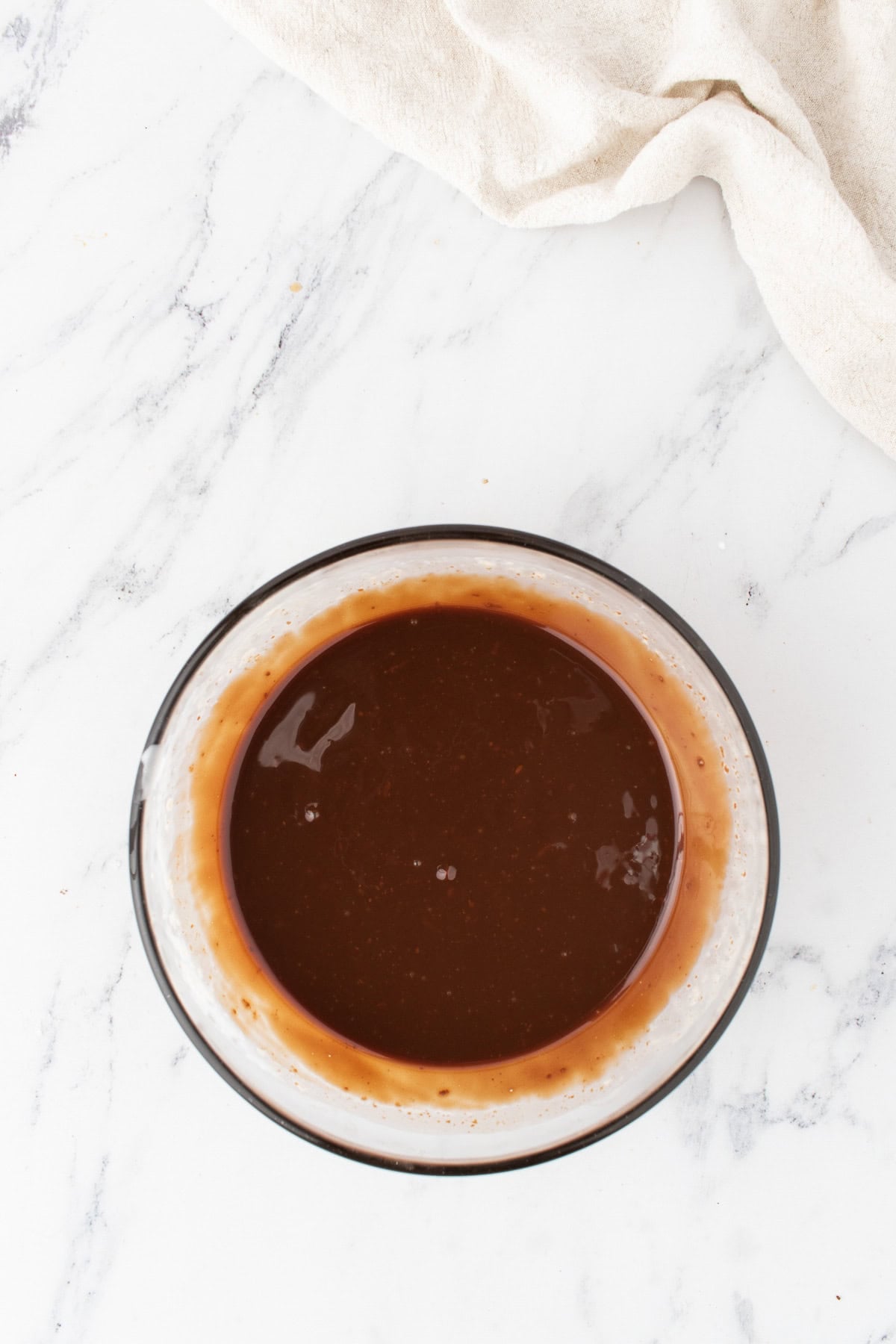 A glass bowl filled with a dark brown liquid sauce, perfect for drizzling over Peanut Butter Chocolate Cake, sits on a white marble surface next to a beige cloth.