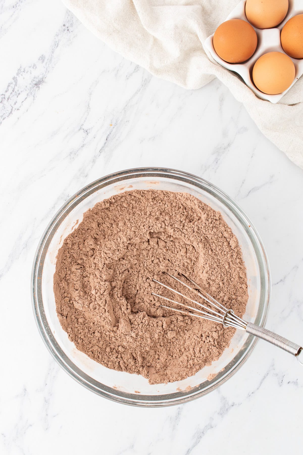 A glass bowl of dry peanut butter chocolate cake mix with a whisk inside, set on a marble surface next to a carton of eggs and a light-colored cloth.