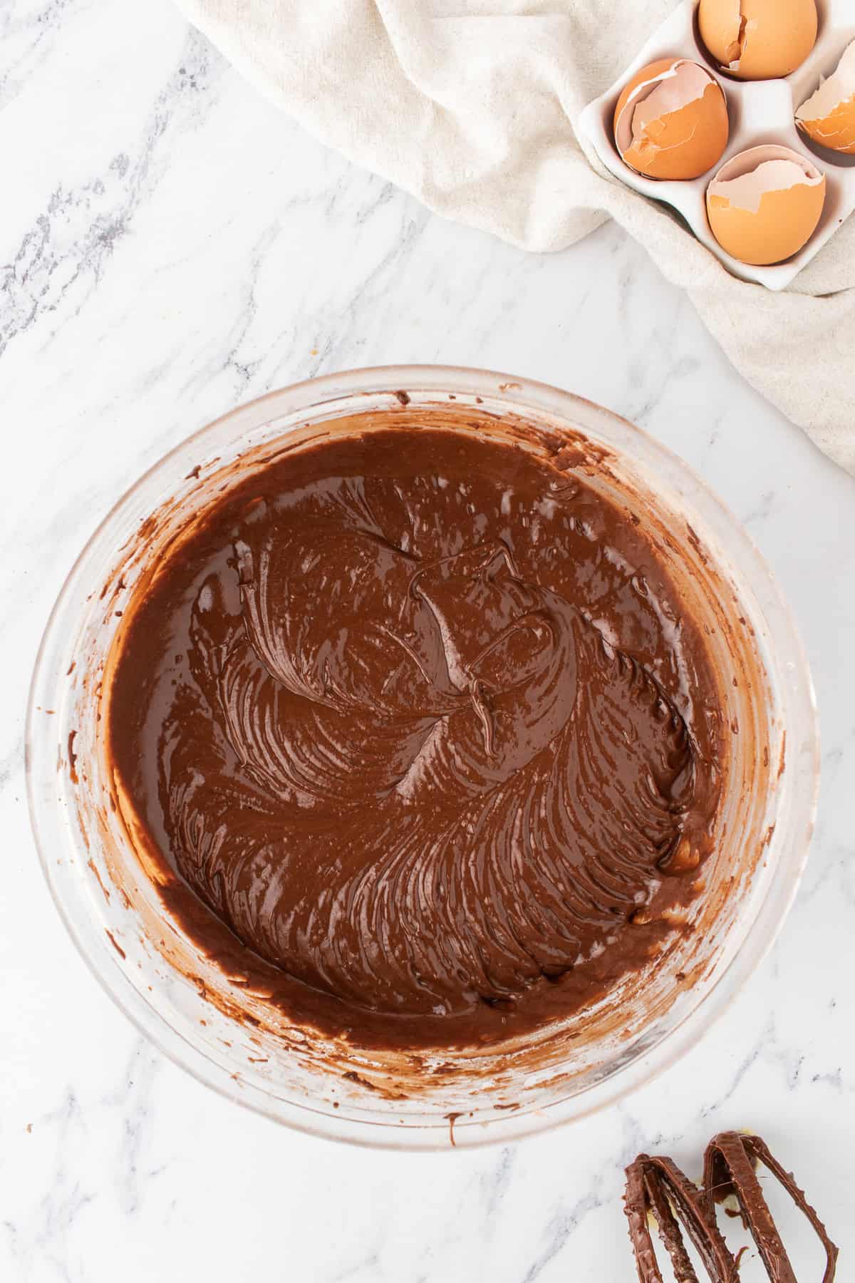 A glass bowl filled with chocolate cake batter sits on a marble surface next to a carton of cracked eggs, creamy peanut butter, and a cloth napkin.