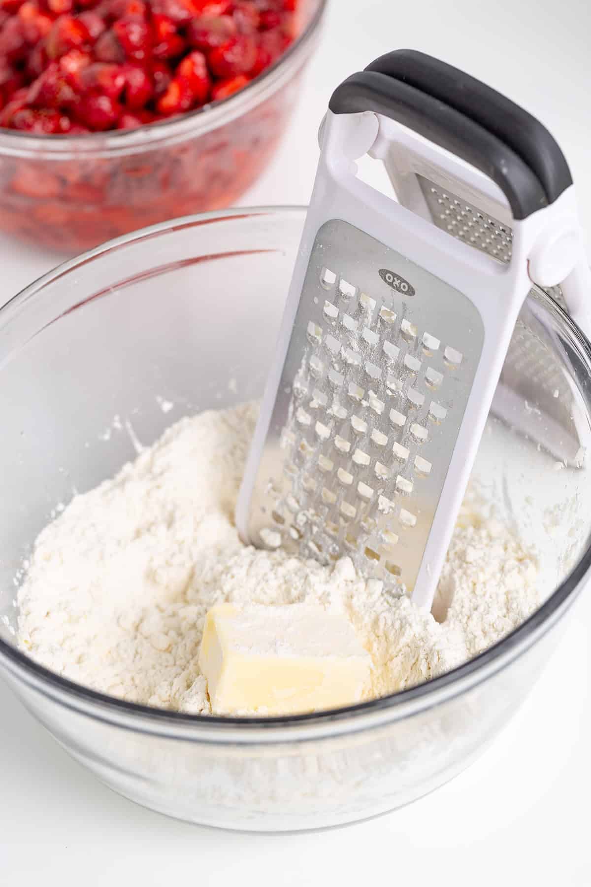 A grater rests in a glass bowl with flour and a chunk of butter, ready for a delicious Strawberry Cobbler; a bowl of chopped strawberries sits in the background.