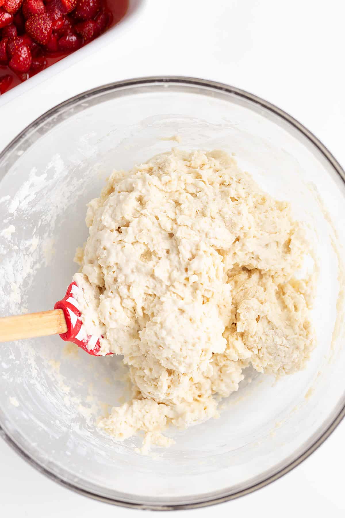 A glass bowl containing biscuit dough being mixed with a red spatula, perfect for a strawberry cobbler recipe, with a dish of sliced strawberries in the background.