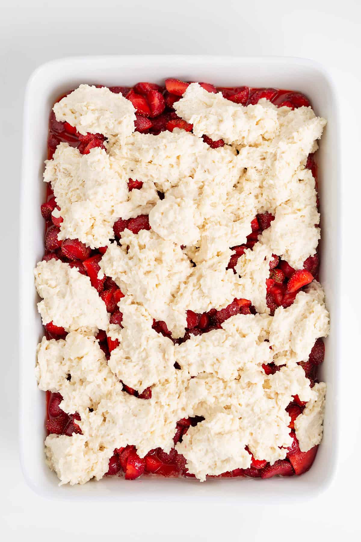 A baking dish filled with sliced strawberries, topped with uneven dollops of biscuit dough, ready to be baked into a delicious strawberry cobbler.