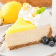A slice of Lemon Cheesecake topped with whipped cream, fresh mint, a lemon garnish, and juicy blueberries on a plate.