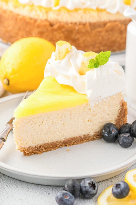 A slice of Lemon Cheesecake topped with whipped cream, fresh mint, a lemon garnish, and juicy blueberries on a plate.