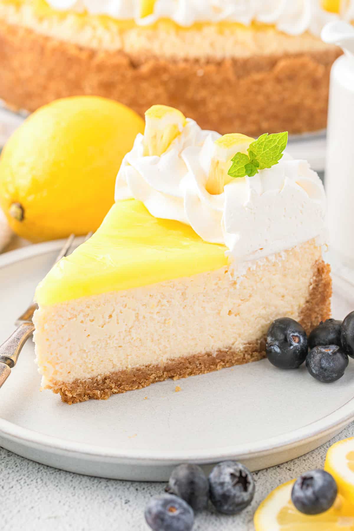 A slice of Lemon Cheesecake topped with whipped cream, fresh mint, a lemon garnish, and juicy blueberries on a plate.