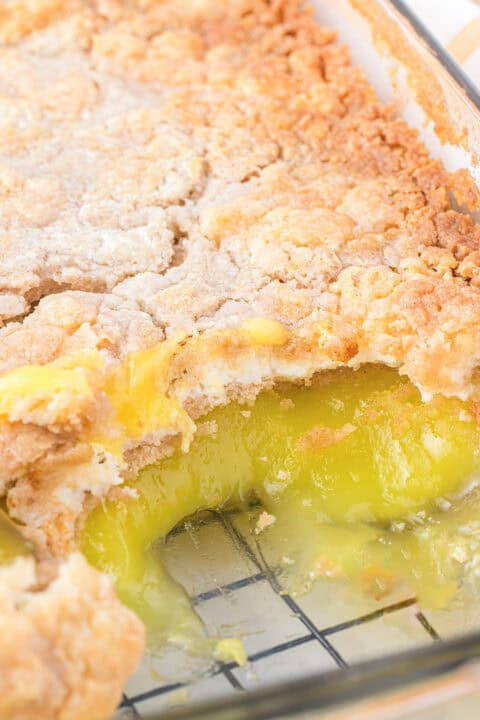 A close-up of a lemon dump cake in a glass dish, featuring a crumbly topping and gooey yellow filling, partially scooped out to reveal its luscious layers.