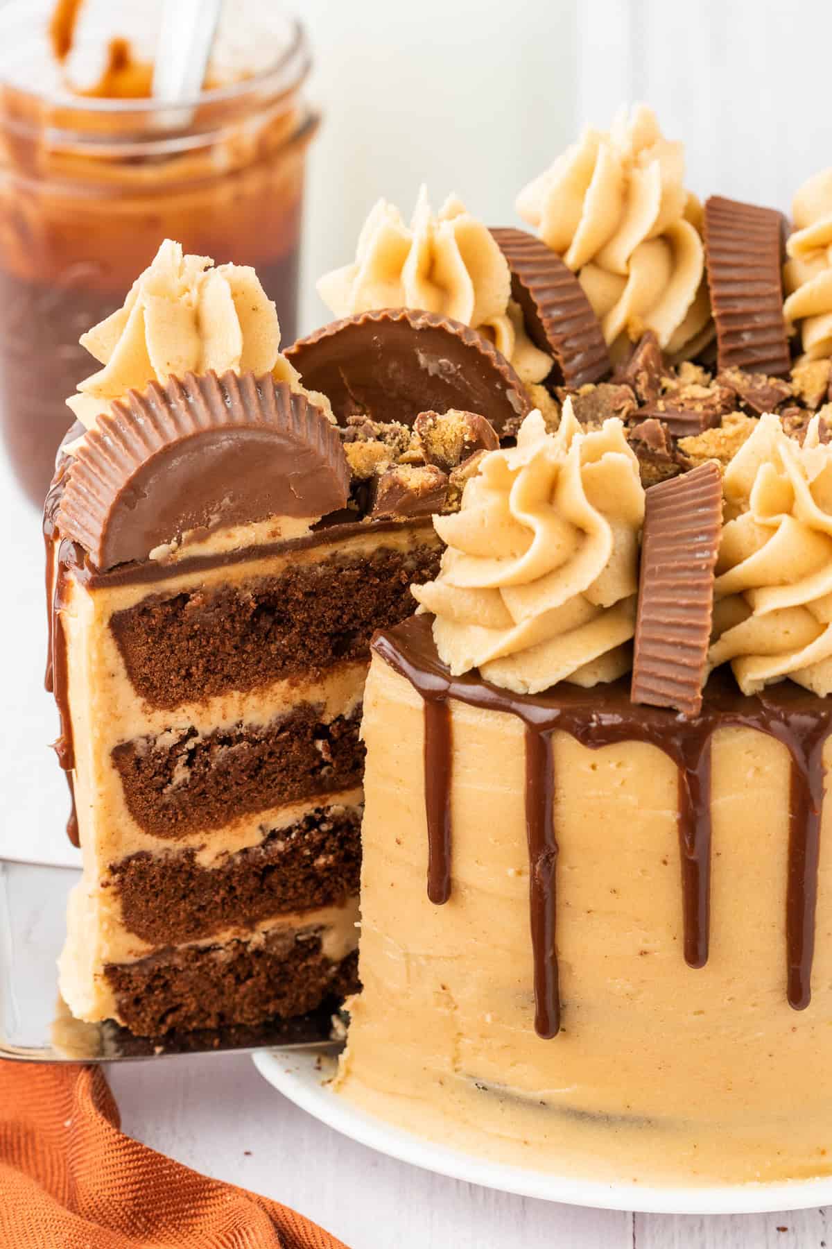 A layered chocolate cake with peanut butter frosting, chocolate drizzle, and peanut butter cup pieces&mdash;a decadent peanut butter chocolate treat, with a slice being served.