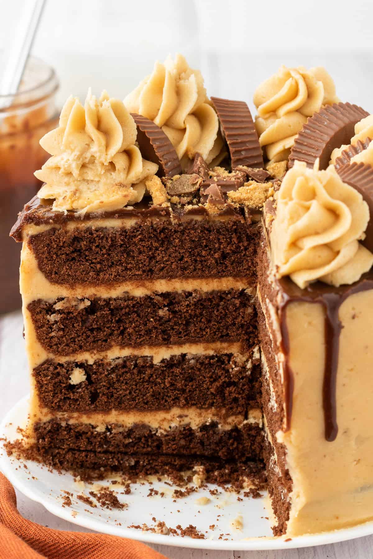 A decadent four-layer peanut butter chocolate cake with peanut butter frosting, topped with peanut butter cups and frosting swirls, sits on a white plate with a slice removed.