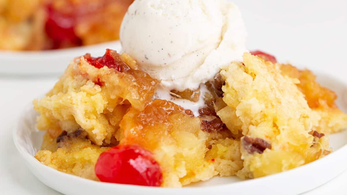 A slice of pineapple upside-down cake topped with vanilla ice cream, featuring visible cherries and cake crumbs on a white plate&mdash;a perfect choice for those craving simple desserts.