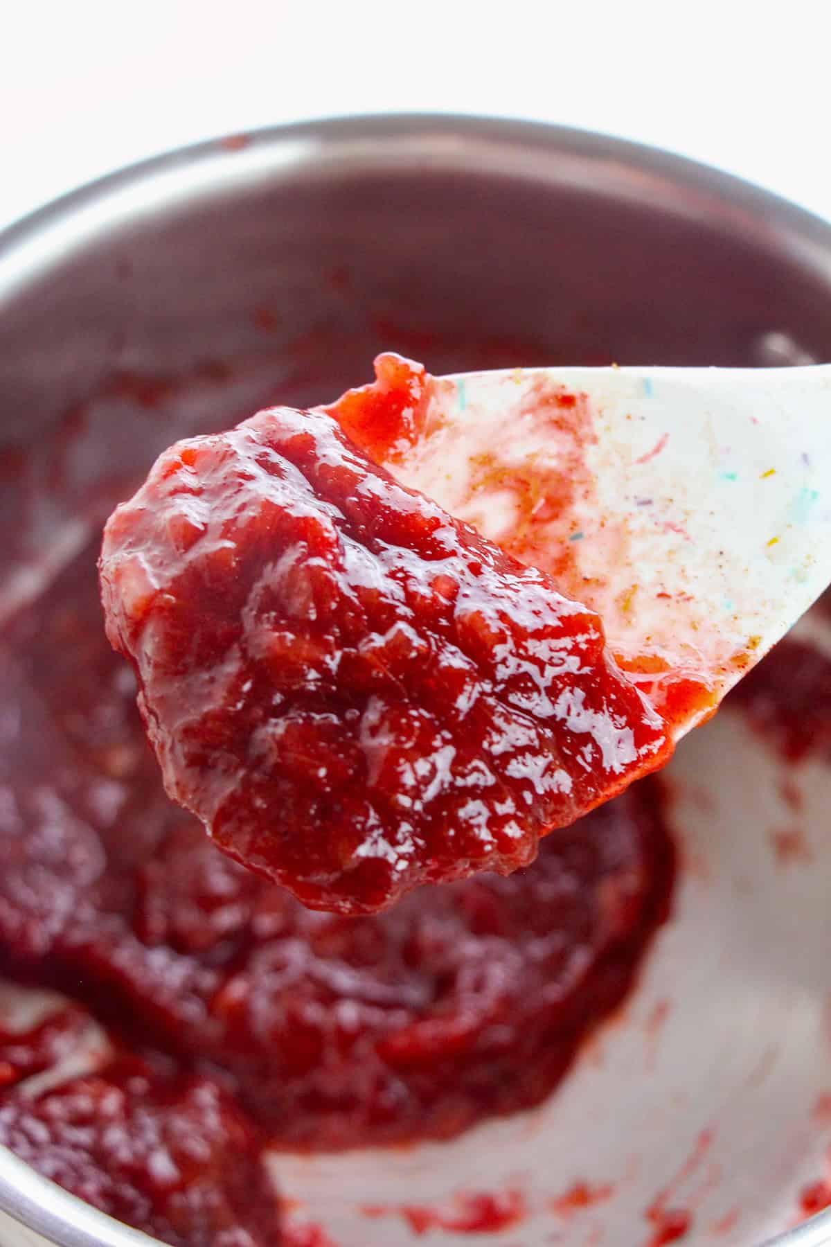 A close-up of a white spoon holding thick, glossy red stewed rhubarb jam above a pot containing more of the same jam&mdash;a delicious rhubarb recipe in the making.