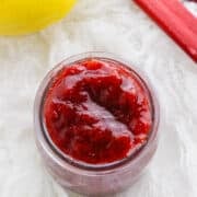 A jar of red fruit jam sits on a white textured surface, with a lemon and stalks of rhubarb nearby—perfect inspiration for your next stewed rhubarb recipe.