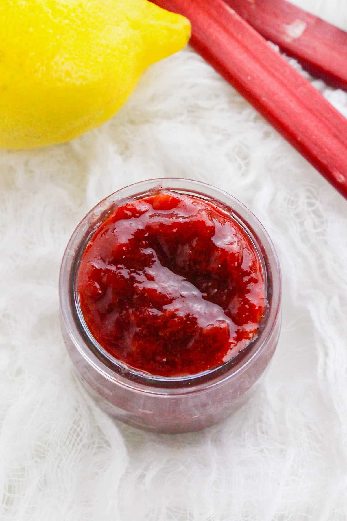 A jar of red fruit jam sits on a white textured surface, with a lemon and stalks of rhubarb nearby&mdash;perfect inspiration for your next stewed rhubarb recipe.