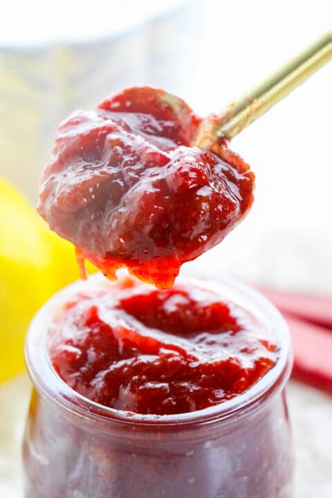 A spoonful of thick, red strawberry jam&mdash;reminiscent of a classic stewed rhubarb recipe&mdash;is held over an open glass jar filled with more delicious jam.