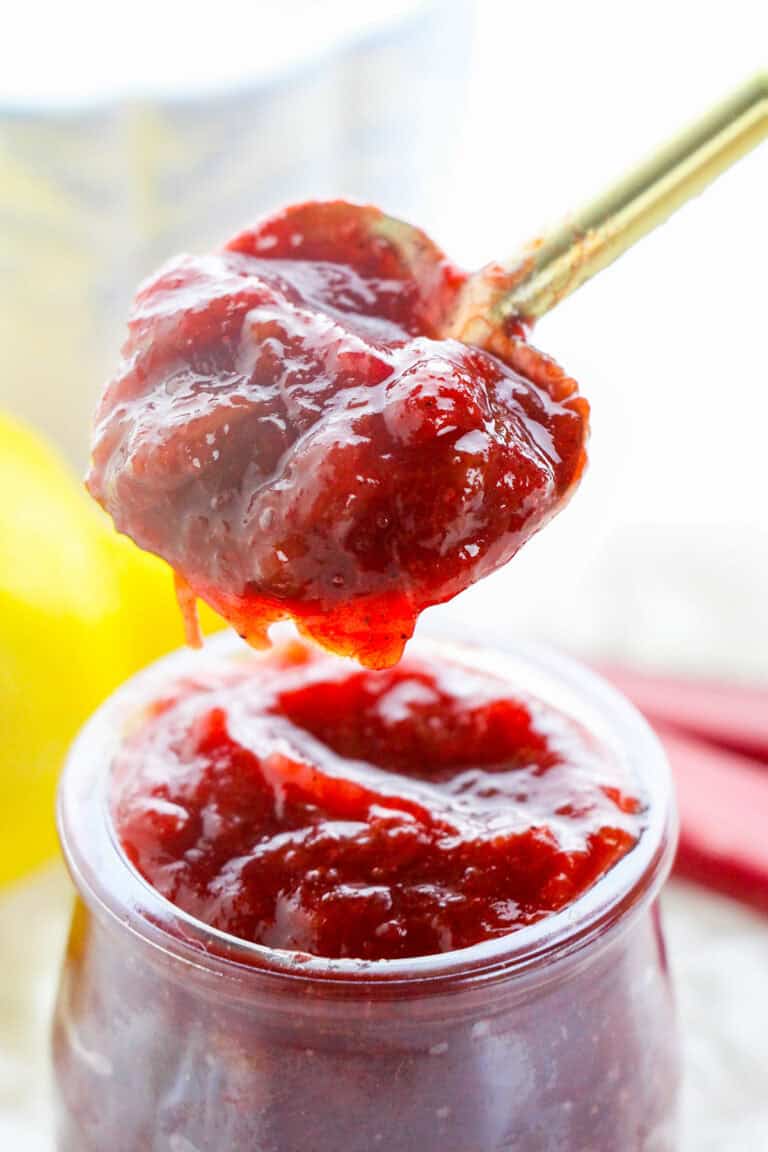 A spoonful of thick, red strawberry jam—reminiscent of a classic stewed rhubarb recipe—is held over an open glass jar filled with more delicious jam.