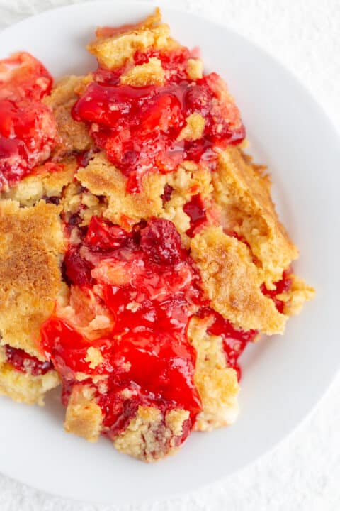 A serving of cherry dump cake with a golden, crumbly topping and bright red cherry filling, perfect for pairing with sweet pineapple or strawberry flavors, on a white plate.