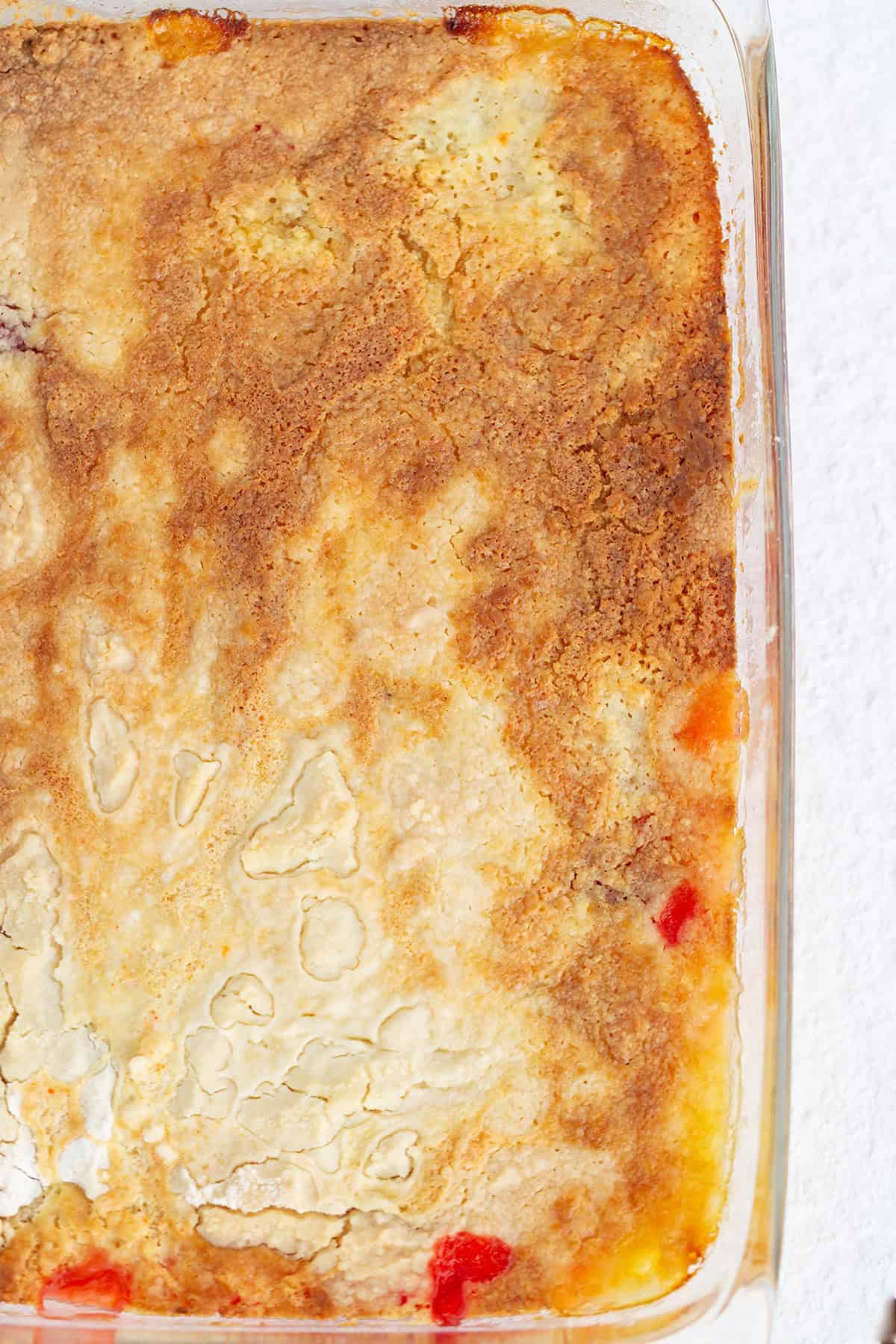 A close-up of a baked peach cobbler in a glass dish, showing a golden-brown, slightly cracked crust with hints of peach filling at the edges&mdash;reminiscent of the tempting layers in a strawberry pineapple dump cake.