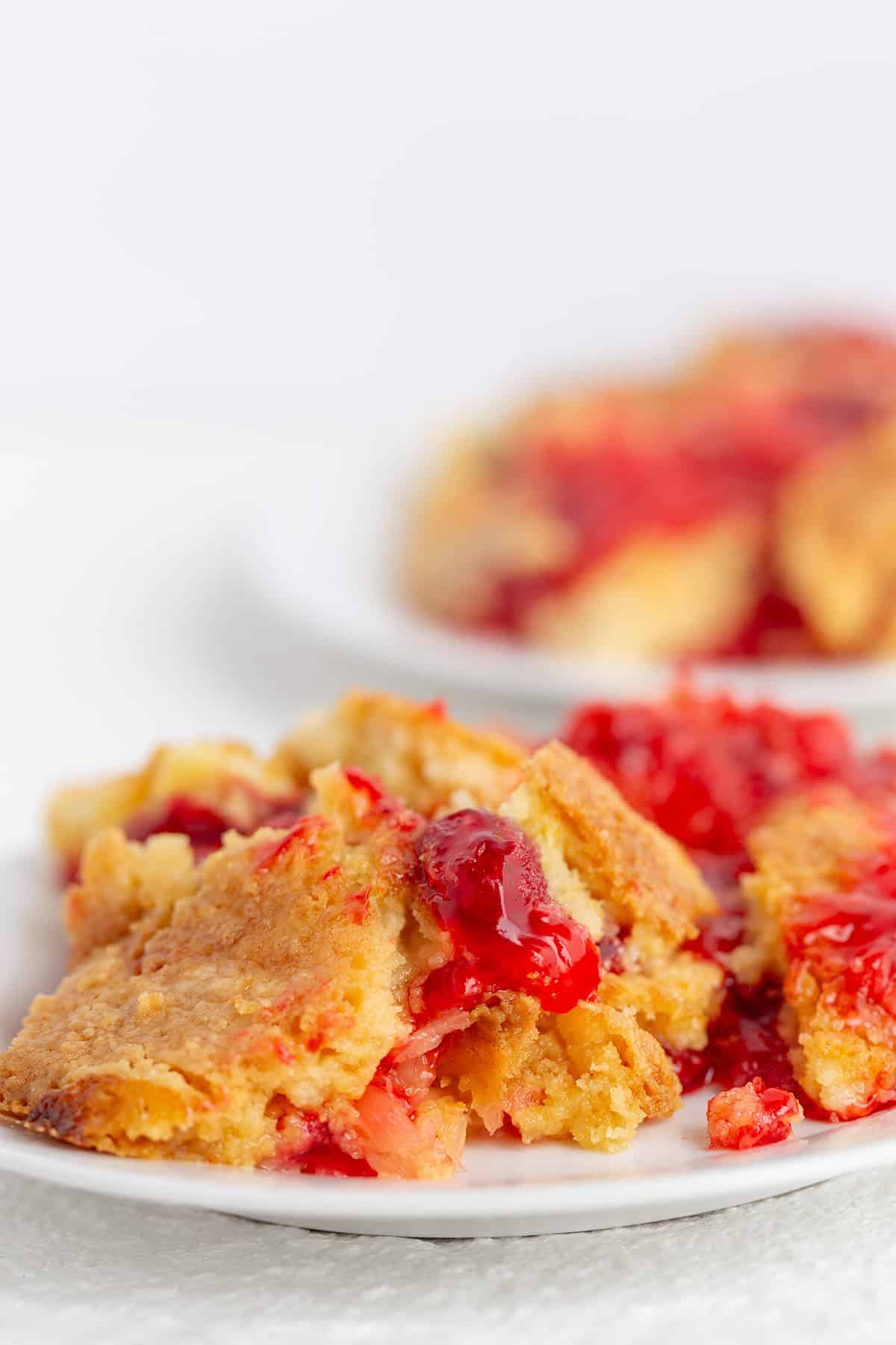 A close-up of a plate with a serving of cherry dump cake, showing a golden crust and bright red cherry filling, hints of pineapple mixed in, with another plate in the background.