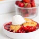 A bowl of Strawberry Cobbler topped with a scoop of vanilla ice cream, with fresh strawberries in a container in the background—a classic strawberry dessert you’ll love.