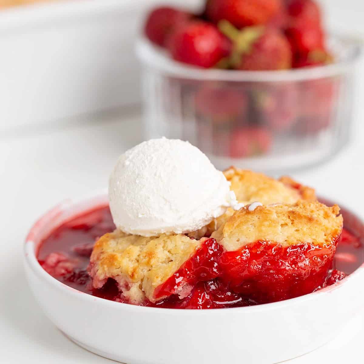 A bowl of Strawberry Cobbler topped with a scoop of vanilla ice cream, with fresh strawberries in a container in the background—a classic strawberry dessert you’ll love.