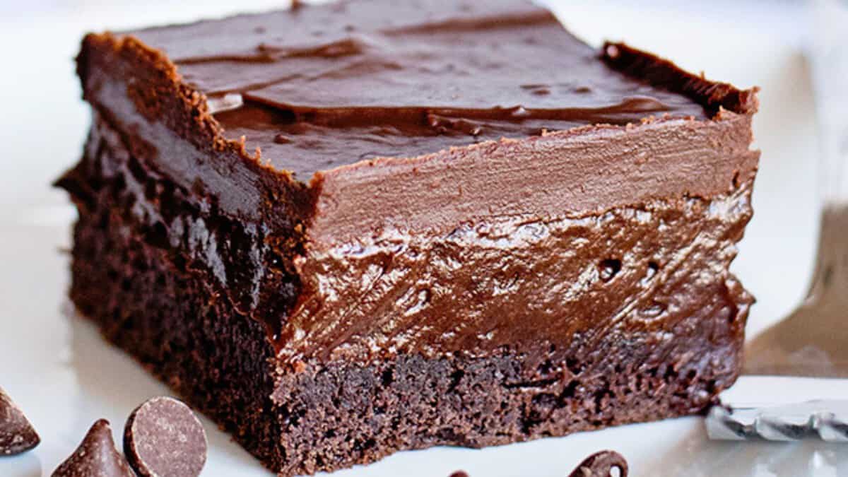 A close-up of a chocolate brownie topped with a thick layer of chocolate frosting, displayed on a plate with chocolate chips nearby&mdash;one of those easy desserts perfect for a stress-free treat.