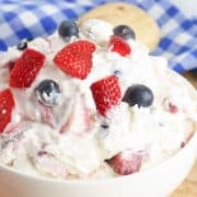 A bowl of Berry Cheesecake Salad with strawberries, blueberries, and whipped cream sits on a wooden surface with a blue and white checkered cloth in the background.