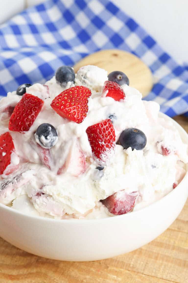 A bowl of Berry Cheesecake Salad with strawberries, blueberries, and whipped cream sits on a wooden surface with a blue and white checkered cloth in the background.